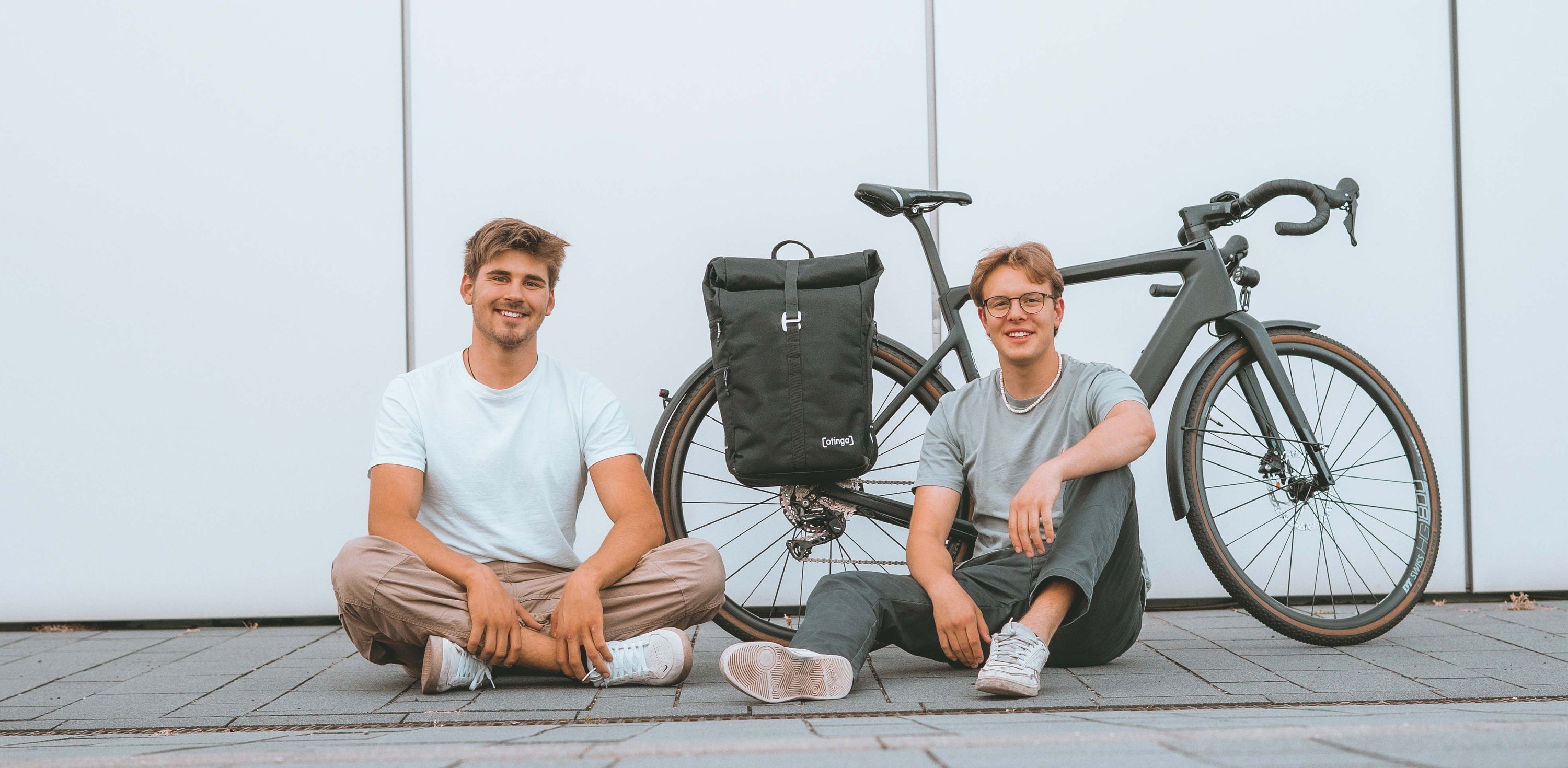 Leander Mellies und Karl Fischer (Aachen, Berlin) die Gründe von otinga / 2bag mit dem Flip / 2bag Rucksack und Fahrradtaschen Kombination auf dem Boden sitzend vor einem Fahrrad